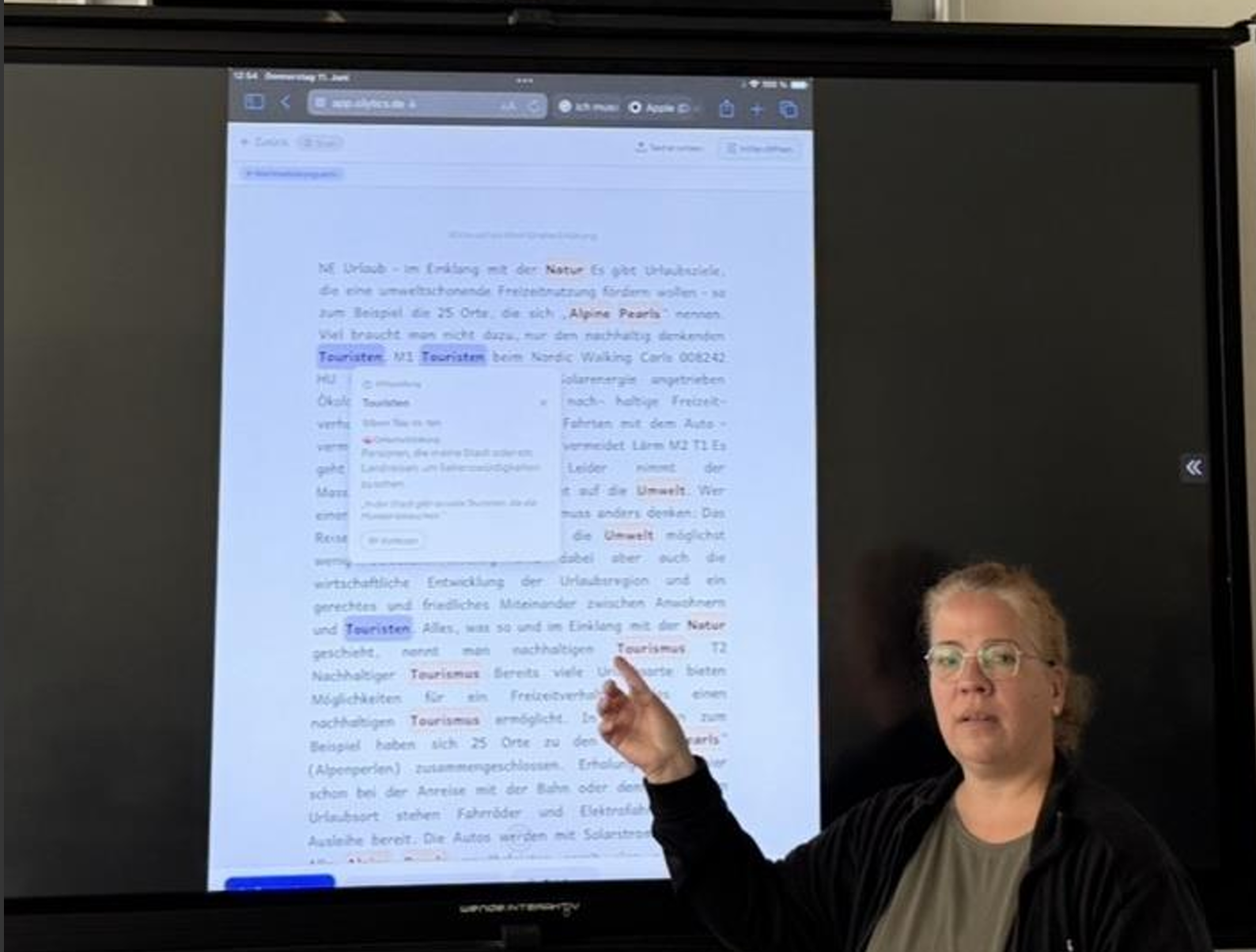 Sandra steht im Klassenzimmer und zeigt auf einen großen Bildschirm mit Silytics — der App-Text mit silbierter Lese-Hilfe ist deutlich sichtbar